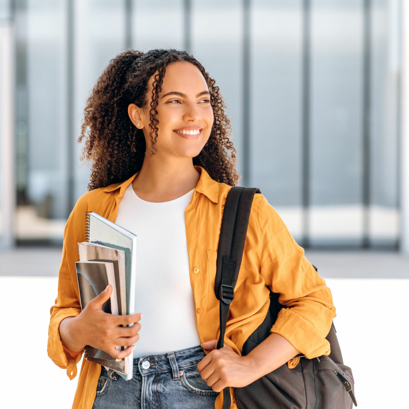 Happy female student. Positive female student, brazilian or hispanic nationality, with a backpack, holding books and notebooks in her hand, stand near the university campus, looks away and smiling